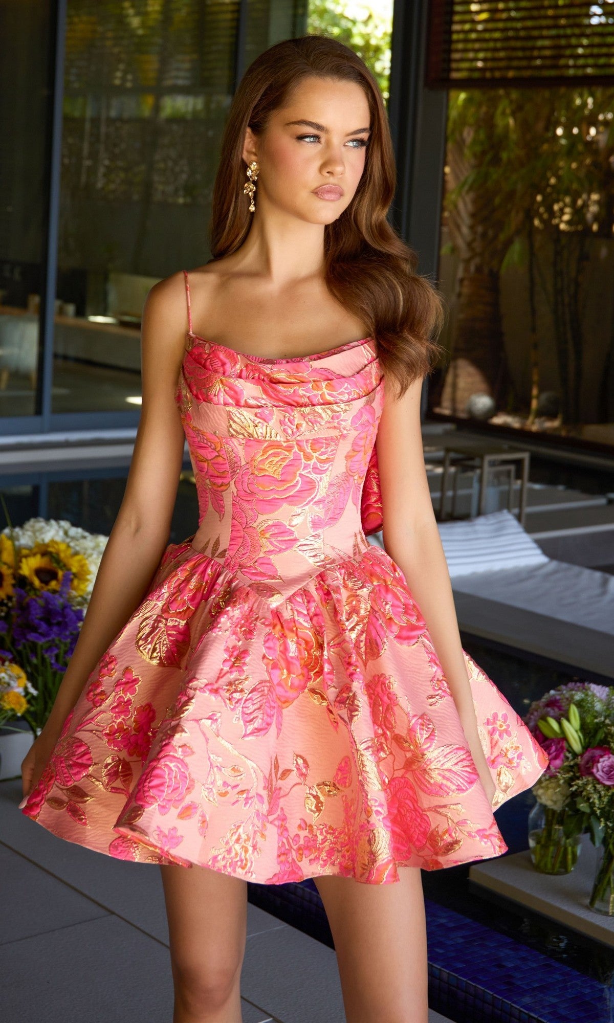 A young woman stands indoors near a glass wall, surrounded by flowers and modern decor, wearing the Ava Presley 42462 short homecoming dress—a sleeveless pink floral jacquard design with a fit-and-flare silhouette.